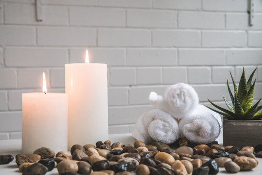 zwei weiße Kerzen mit unterschiedlichen Längen, Kaktuspflanze, 3 Badetücher und vielen kleine Standsteine mit einer weißen Wand im Hintergrund.