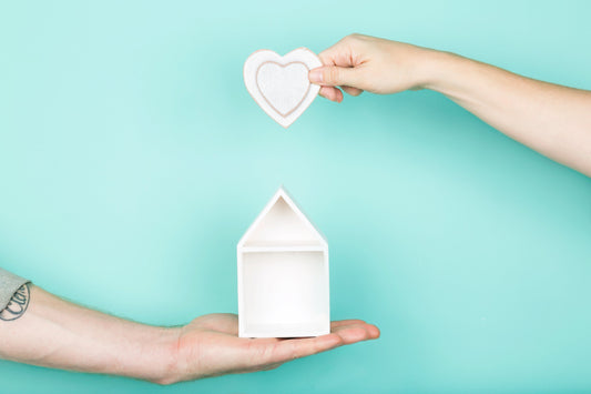 zwei Hände; eine Hand hält ein Herz auf Pappe und die Andere hält ein Haus aus Pappe mit einem türkis Hintergund.