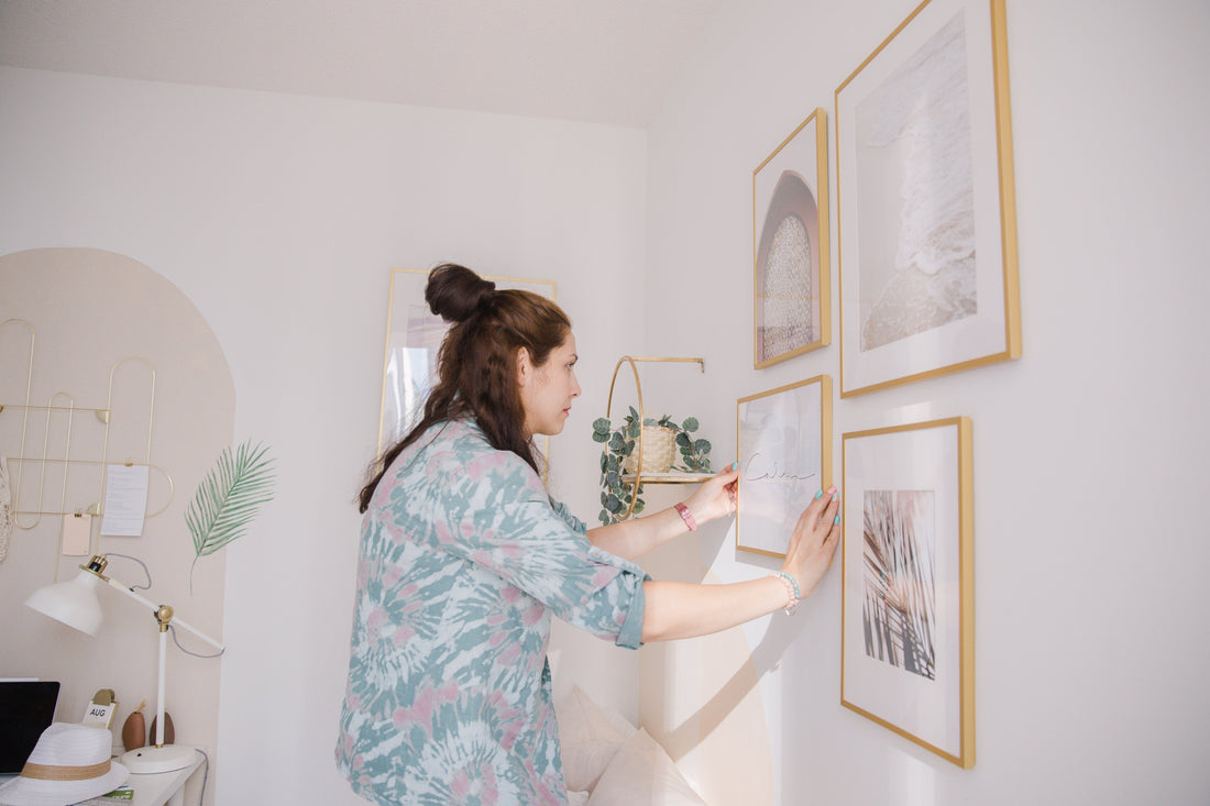 Eine Frau in einem weißen Haus platziert einen golden Bilderrahmen auf die Wand. Da sind auch andere goldene Bilderrahmen daneben auf die Wand mit verschiedenen größen zu sehen