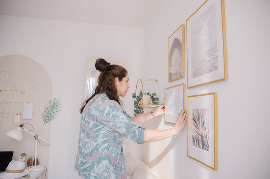 Eine Frau in einem weißen Haus platziert einen golden Bilderrahmen auf die Wand. Da sind auch andere goldene Bilderrahmen daneben auf die Wand mit verschiedenen größen zu sehen
