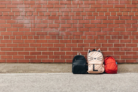 Drei Taschen (schwarze, pinke, rote) mit verschiedenen Größen am Boden auf der rechten Ecke des Bildes mit einem orangen Blockwand im Hintergrund.  